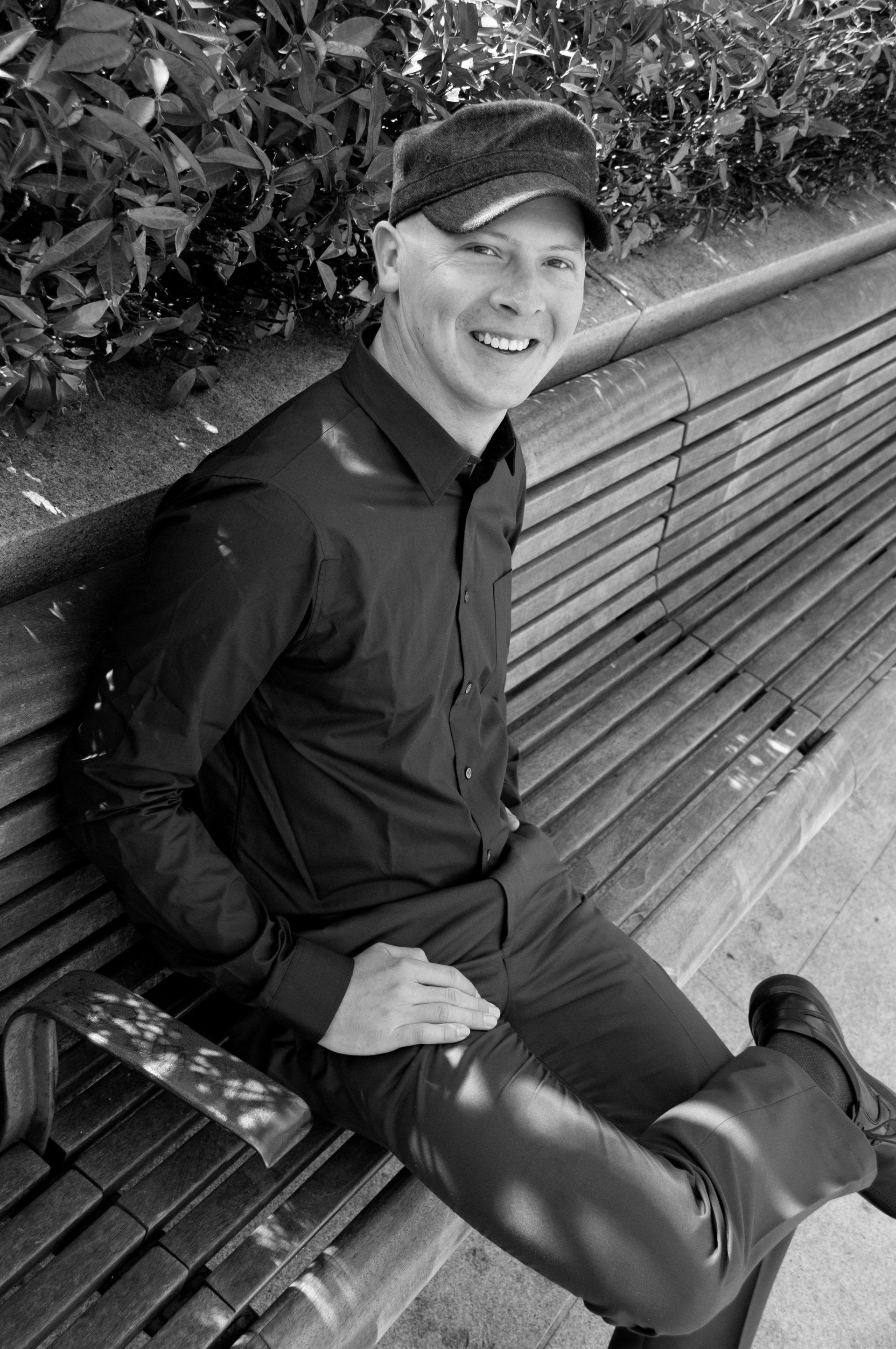 Man smiling on a park bench, wearing dark shirt and cap, sunlight dappling his face.