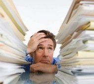 Een gestresste man, omringd door stapels papier, houdt zijn hoofd tegen een bureau.