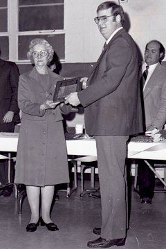 Woman in dress suit receives a framed object from a man in a blazer, indoors. Other people are in the background.