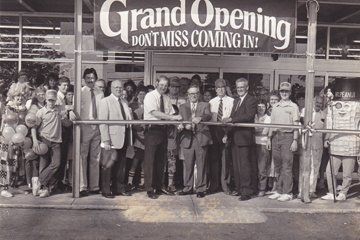 Grand opening ribbon-cutting ceremony at a store, with many people gathered and a banner overhead.