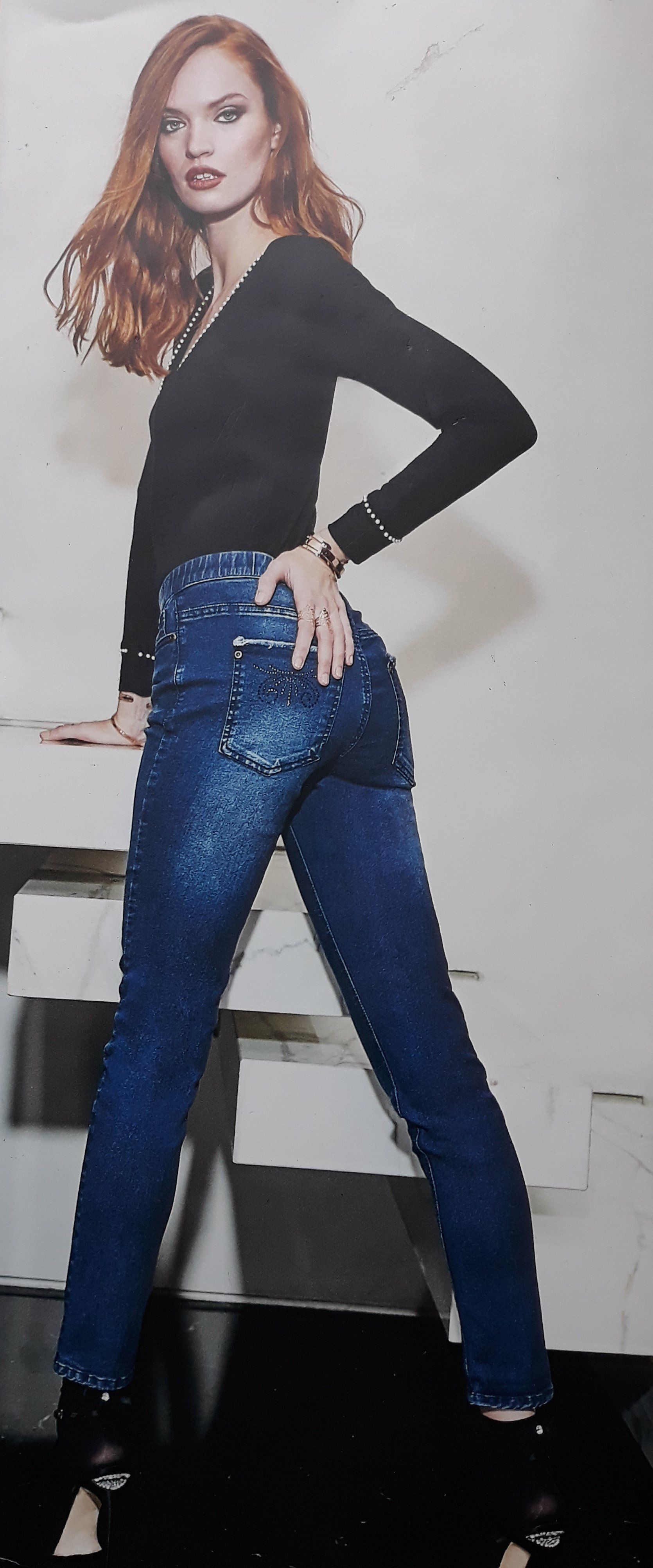 Donna con top nero e jeans blu decorati, in posa su uno sfondo bianco.