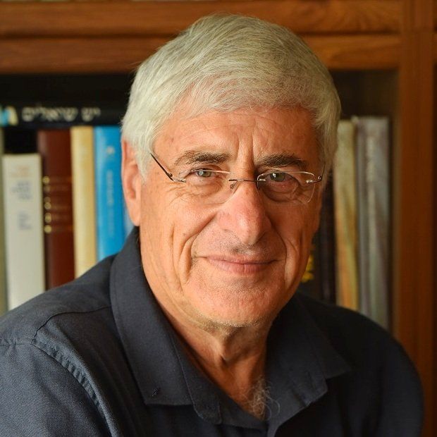 גבר עם שיער אפור ומשקפיים מחייך מול מדף ספרים.