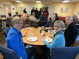 People seated around a banquet table in a community room, smiling and holding drinks.