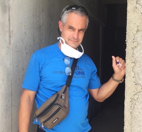 Man in blue shirt standing in a doorway, wearing a neck fan and crossbody bag.