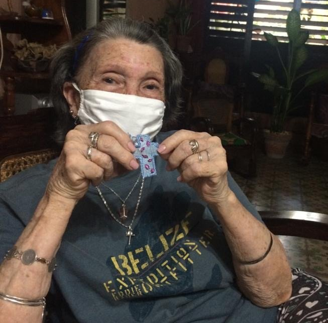 Woman indoors holding a small purple cloth, wearing a white face mask and dark T-shirt, seated in a home setting