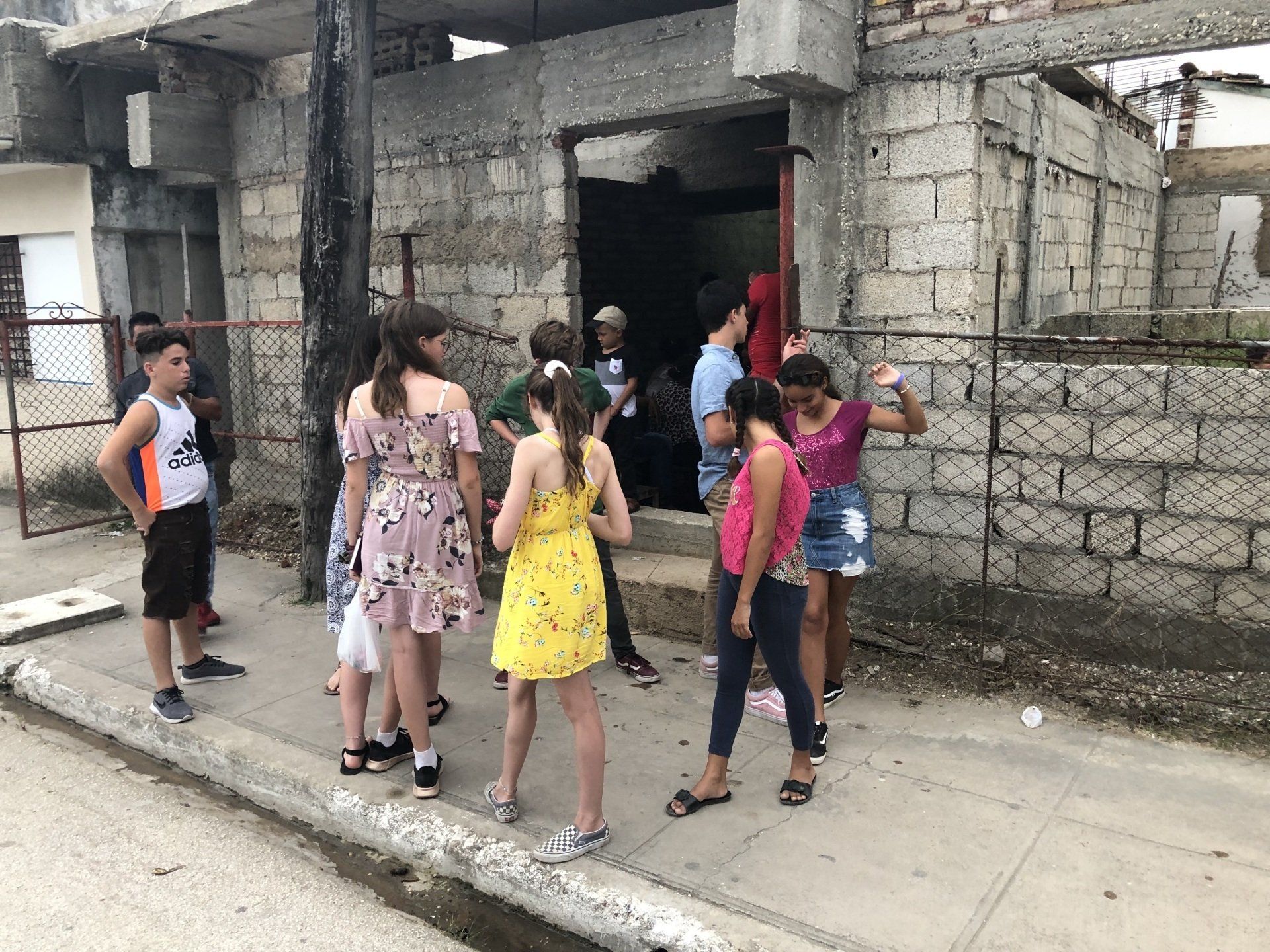 People gathered on a sidewalk beside a concrete building, some talking and standing near an open doorway.