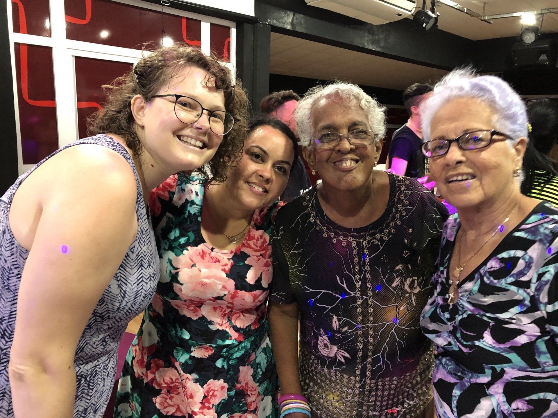 Four smiling people pose together indoors, wearing colorful dresses and glasses.