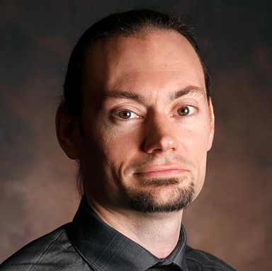 Studio portrait of a person with dark hair and a goatee, wearing a dark shirt against a soft brown backdrop