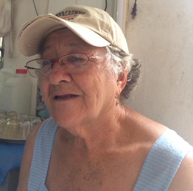 Person wearing a tan cap and glasses, smiling in a sleeveless light blue top outdoors.