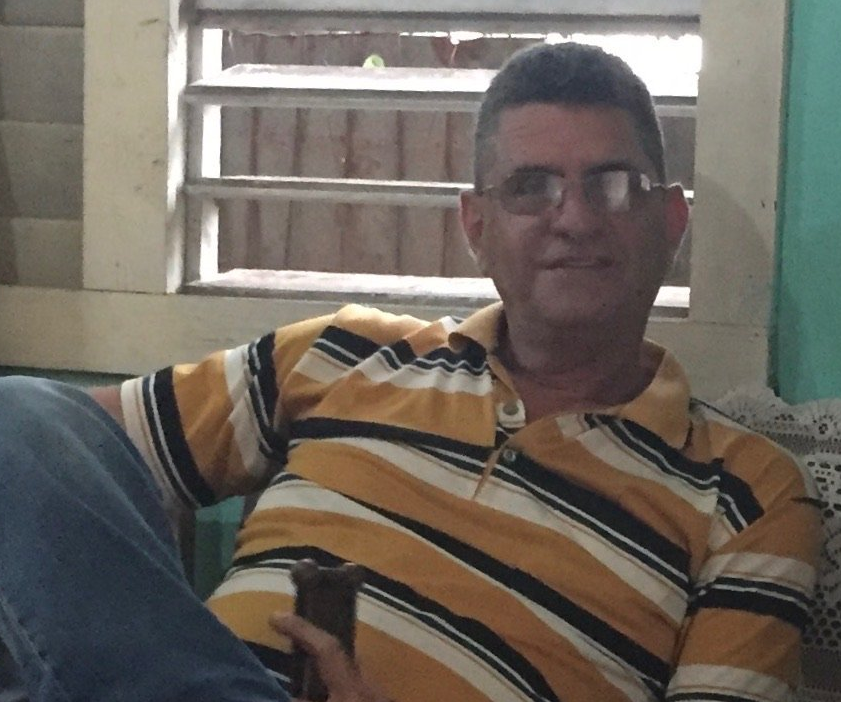 Man seated indoors in a yellow striped polo shirt, wearing glasses, near a window with blinds.
