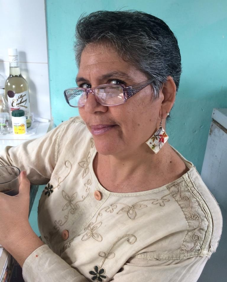 Smiling woman with glasses and earrings, seated indoors against a teal wall.
