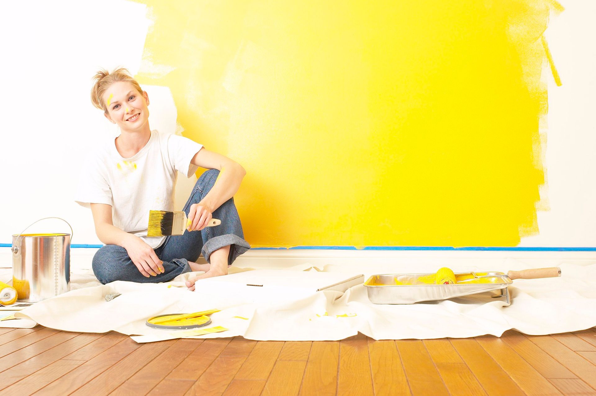 Person sitting on a drop cloth, painting a wall bright yellow with paint cans and supplies nearby