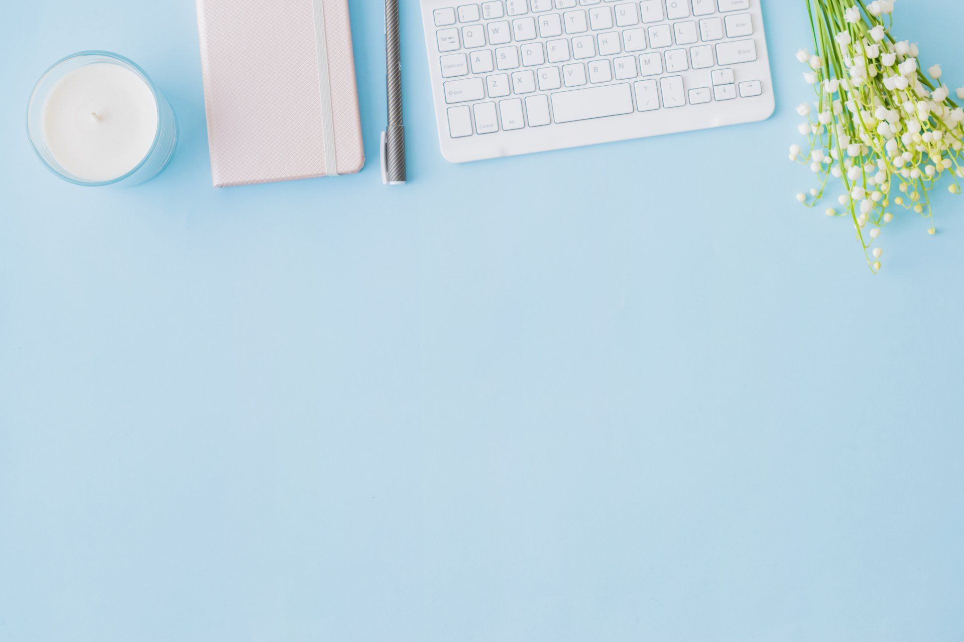 A blue desk with a keyboard , notebook , pen , candle and lily of the valley flowers.