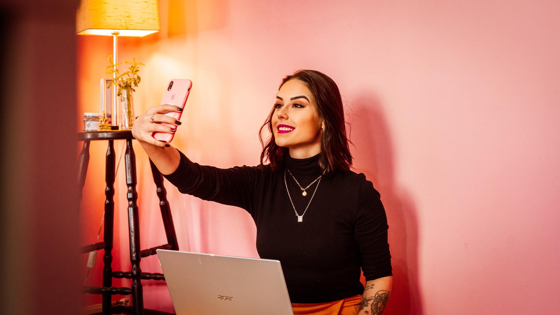 A woman is taking a selfie with her phone while sitting in front of a laptop.
