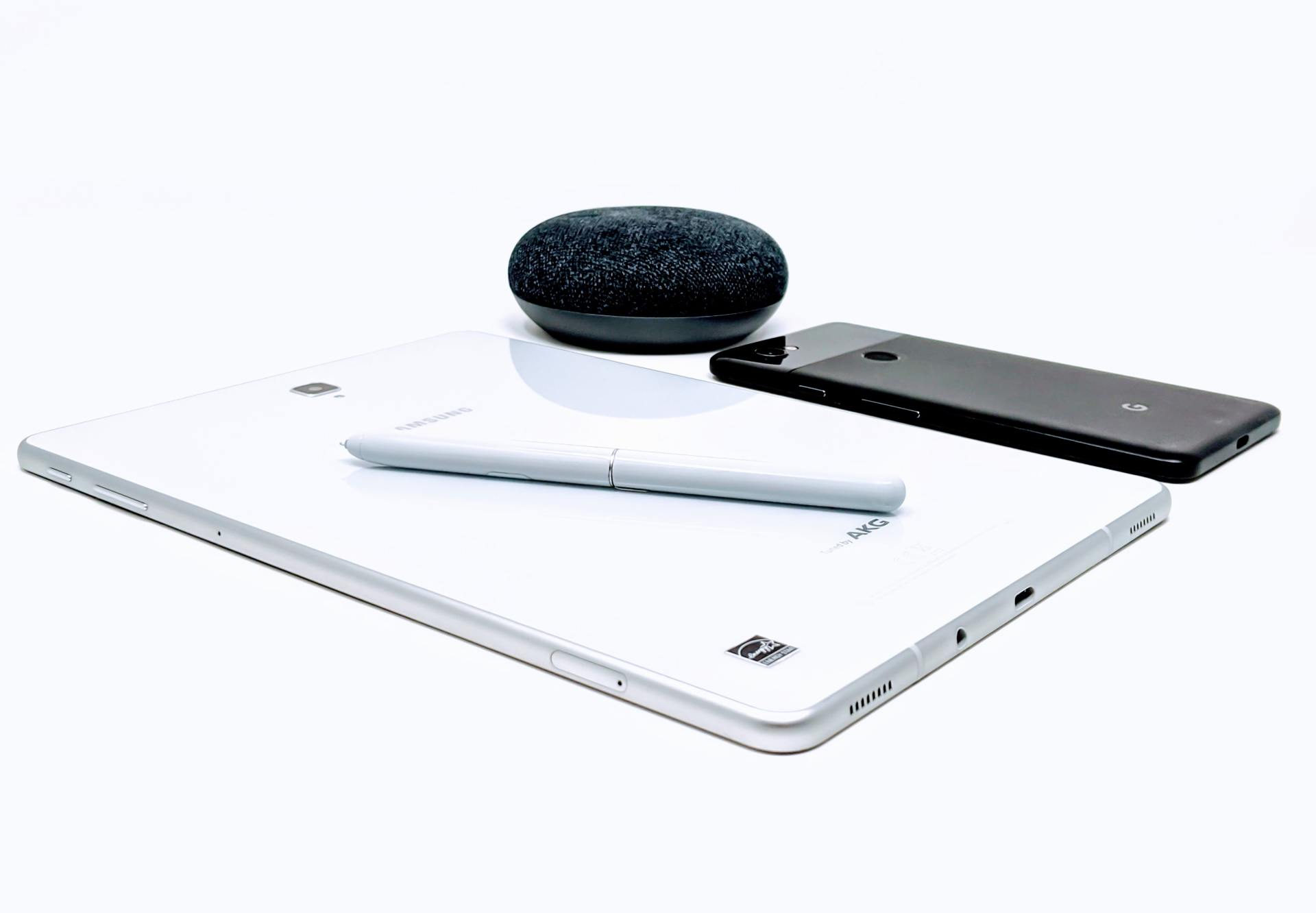 A tablet , phone , and speaker are sitting on top of each other on a white surface.