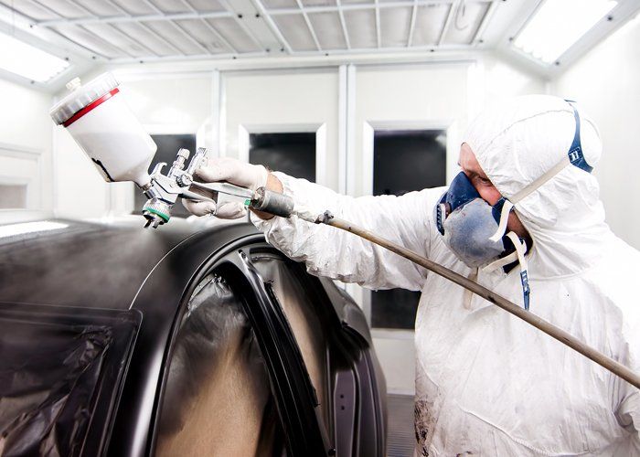 Auto painter spraying a car body with black paint and protective gear