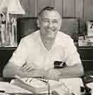 Smiling older man in white shirt, seated at a desk.