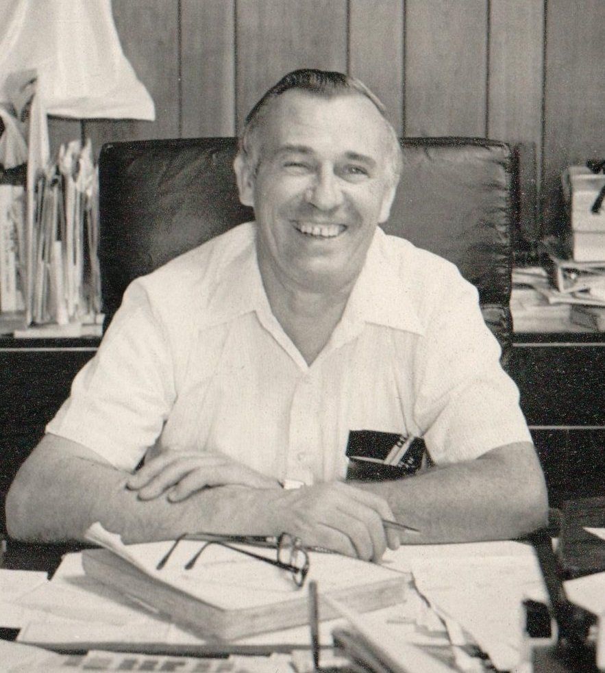 Smiling older man sitting at a desk, hands clasped, wearing a short-sleeved shirt, office setting.