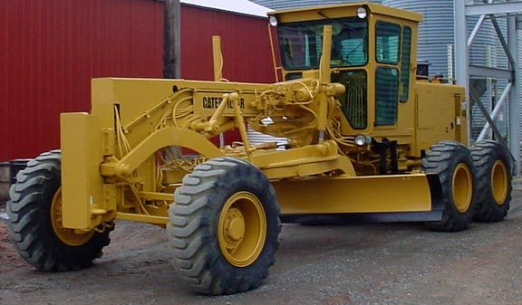 Yellow Caterpillar motor grader parked near a red building and a silo.