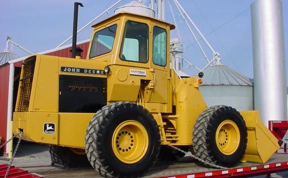 Yellow John Deere wheel loader on a flatbed trailer, in front of silos and a red building.
