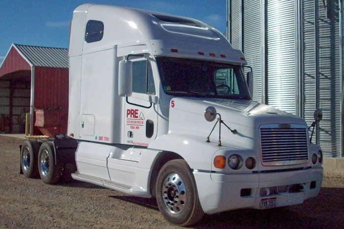 White Freightliner semi-truck parked outdoors next to a metal grain silo and red barn.