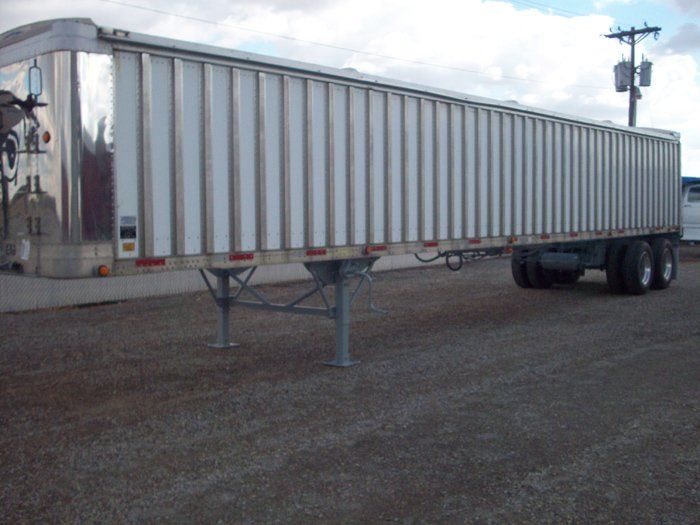 Silver semi-trailer with vertical lines on a gravel surface next to utility pole.