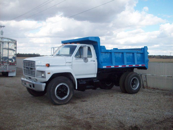 White and blue dump truck parked on gravel. Cloudy sky in the background.