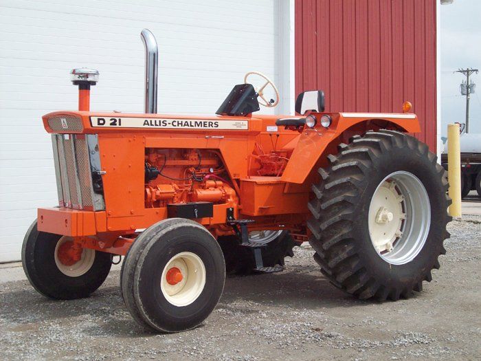 Orange Allis-Chalmers D21 tractor with large rear tires, parked near a red building.