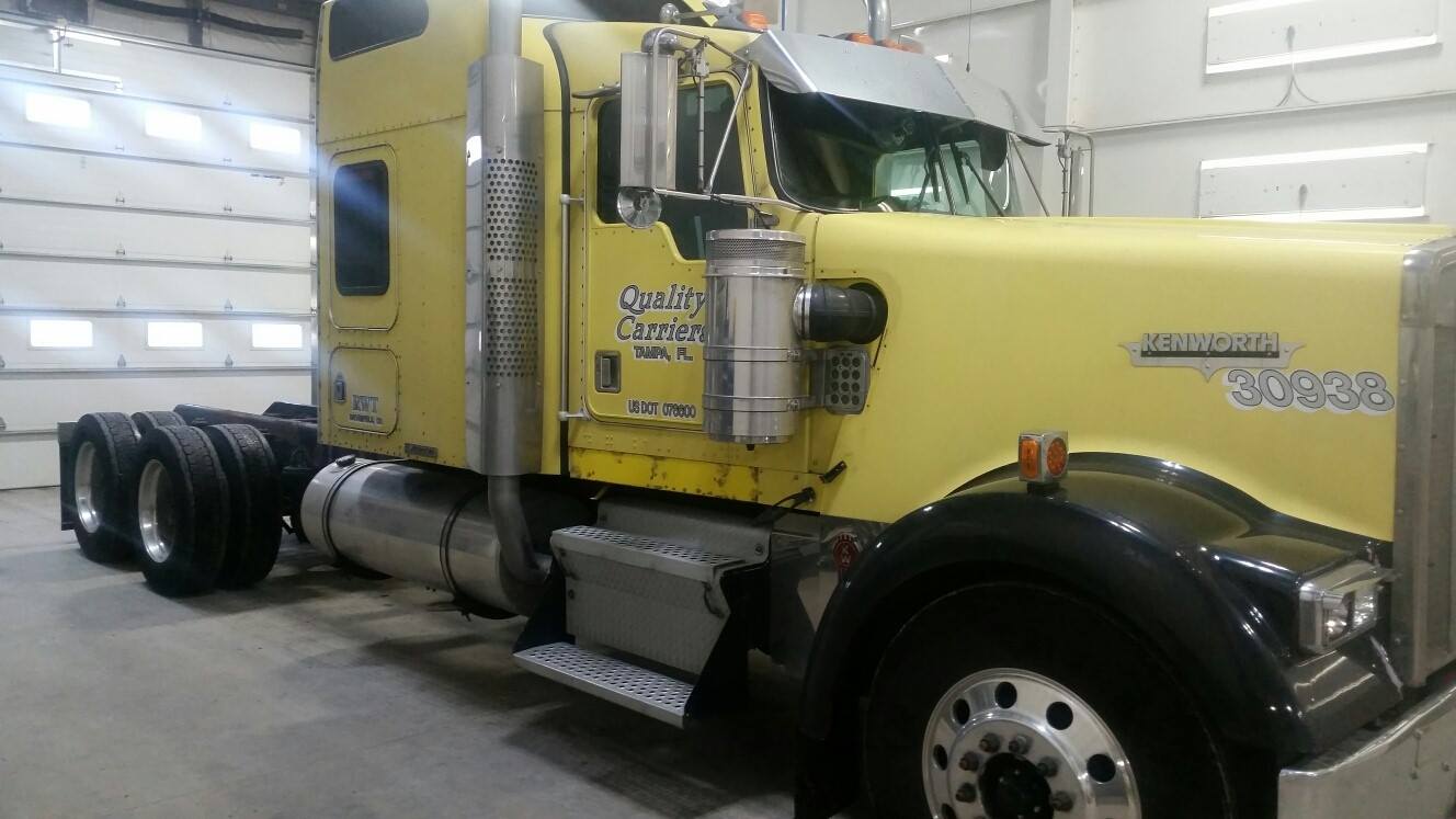 Yellow Kenworth semi-truck in a garage, with silver accents and black fenders, parked in front of a white wall.