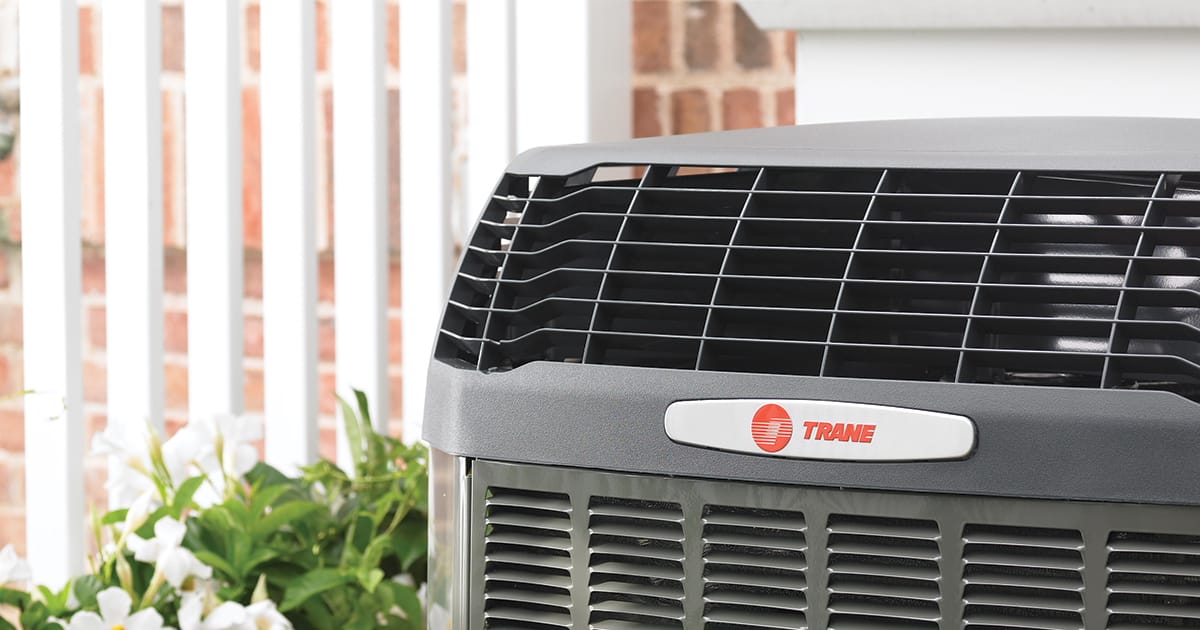 Air conditioning unit with Trane logo, outdoors near white fence and bushes.