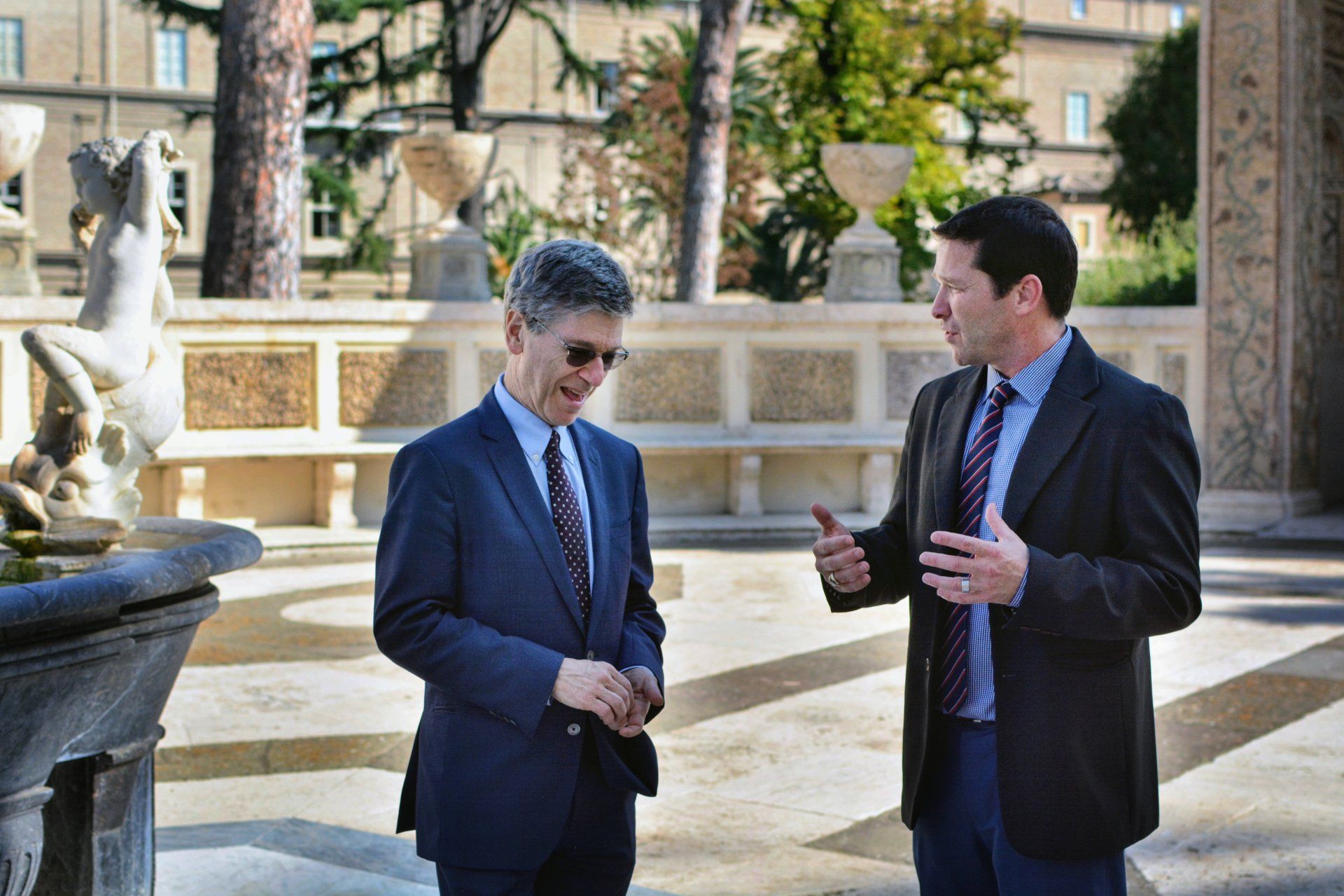 Two men in suits and ties are talking in front of a fountain.