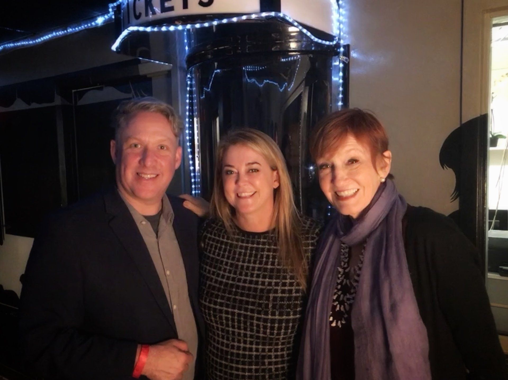 Three smiling people pose together indoors in front of a ticket booth decorated with lights.