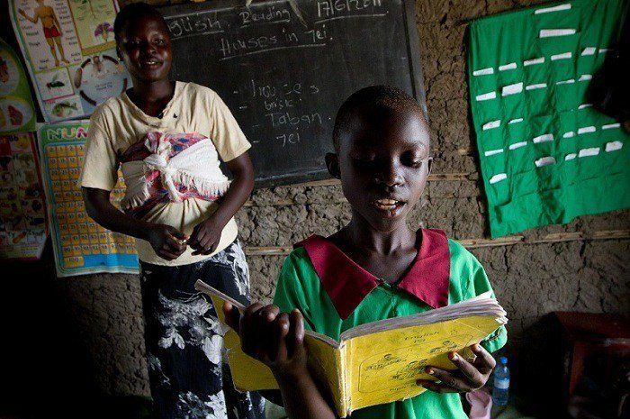 A student reading a book with the teacher standing behind him. 