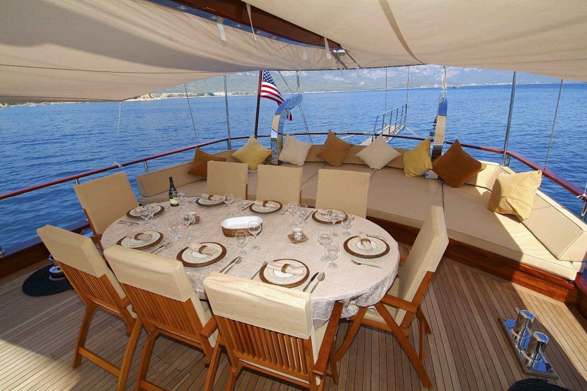 Eten in buitenlucht op een jacht. Een tafel gedekt, omringd door stoelen en comfortabele kussens;  zee op de achtergrond.