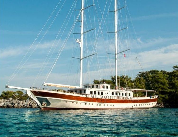 Zeilboot met drie masten op turquoise water, witte romp en houten accenten, vlakbij een rotsachtige kust en bomen.
