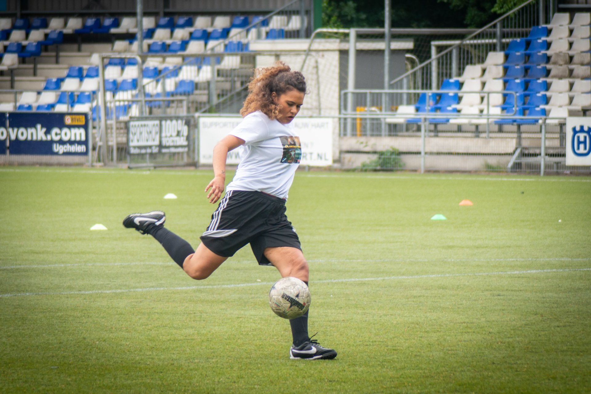 Een persoon in een wit shirt en een zwarte korte broek trapt een voetbal op een groen veld voor de tribune van een stadion.