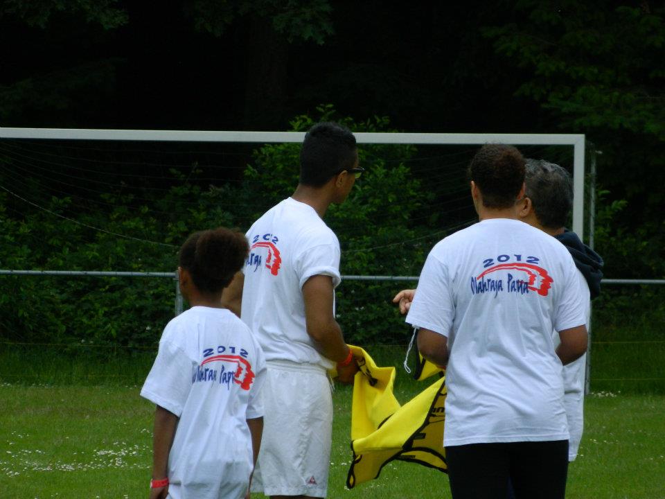 Drie mensen in witte T-shirts met een 2012-logo staan ​​op een veld voor een voetbaldoel en houden een geel shirt vast.