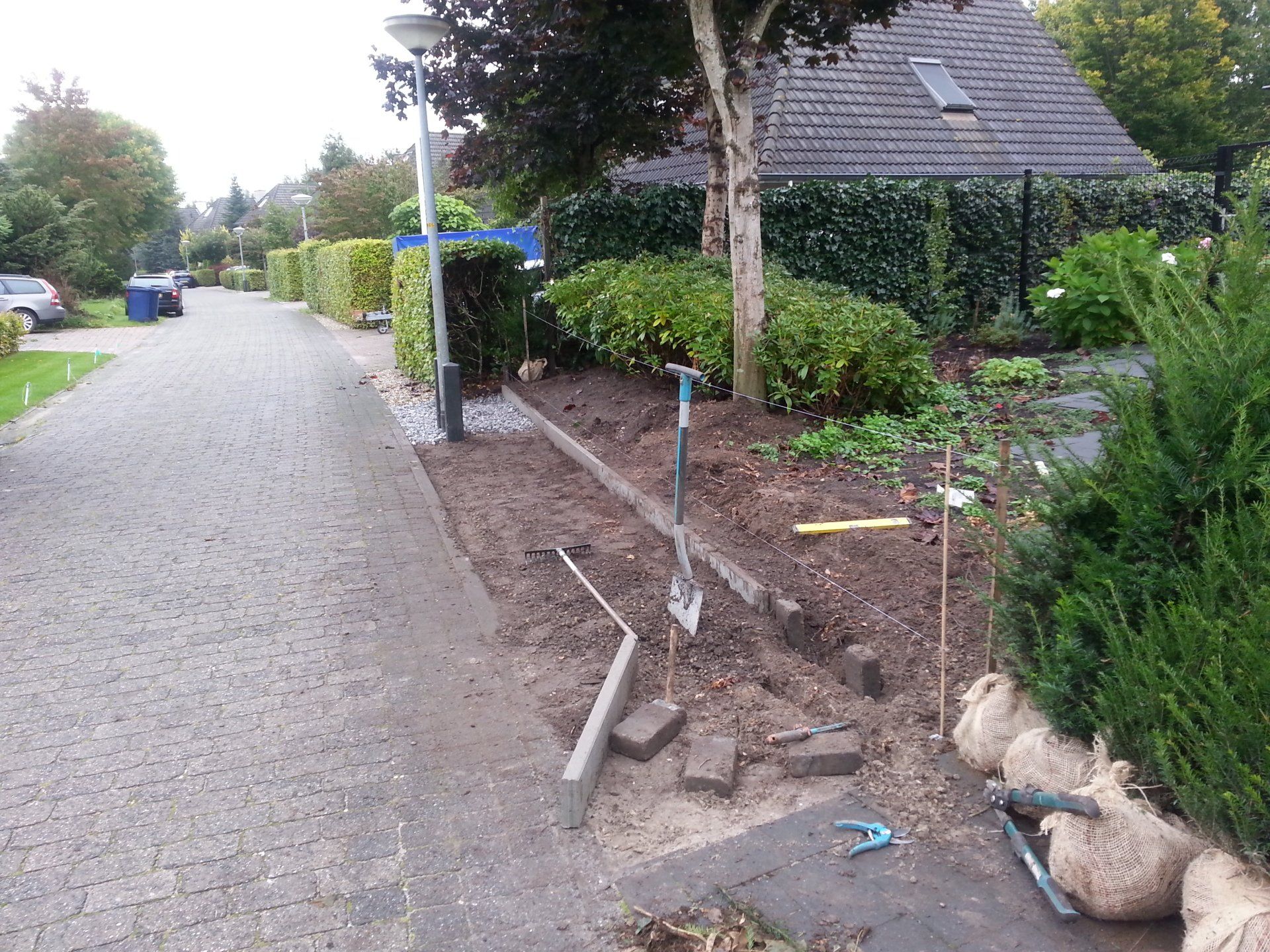 Woonstraat met uitgegraven trottoir, zichtbare leidingen en gestapelde straatstenen naast een heg en een huis.