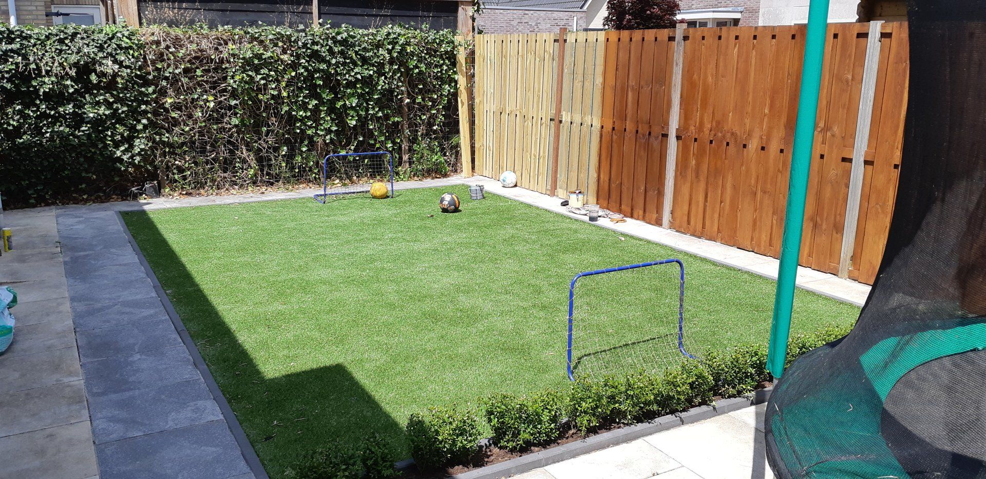 Kleine achtertuin met kunstgras, houten schutting, haag en een blauw frame bij het terras.