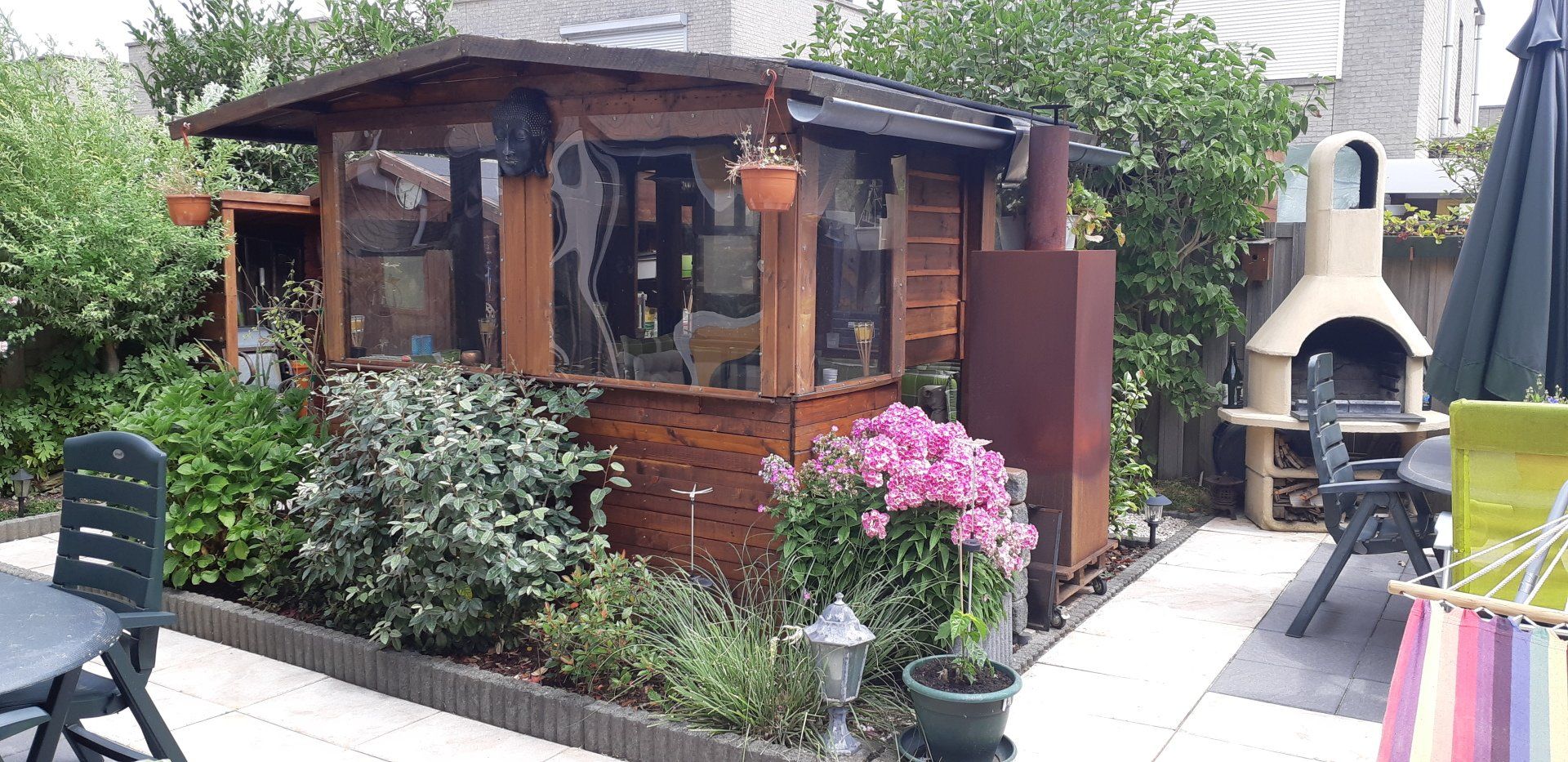 Houten tuinhuisje met ramen, omgeven door bloemen en planten op een terras in de achtertuin.