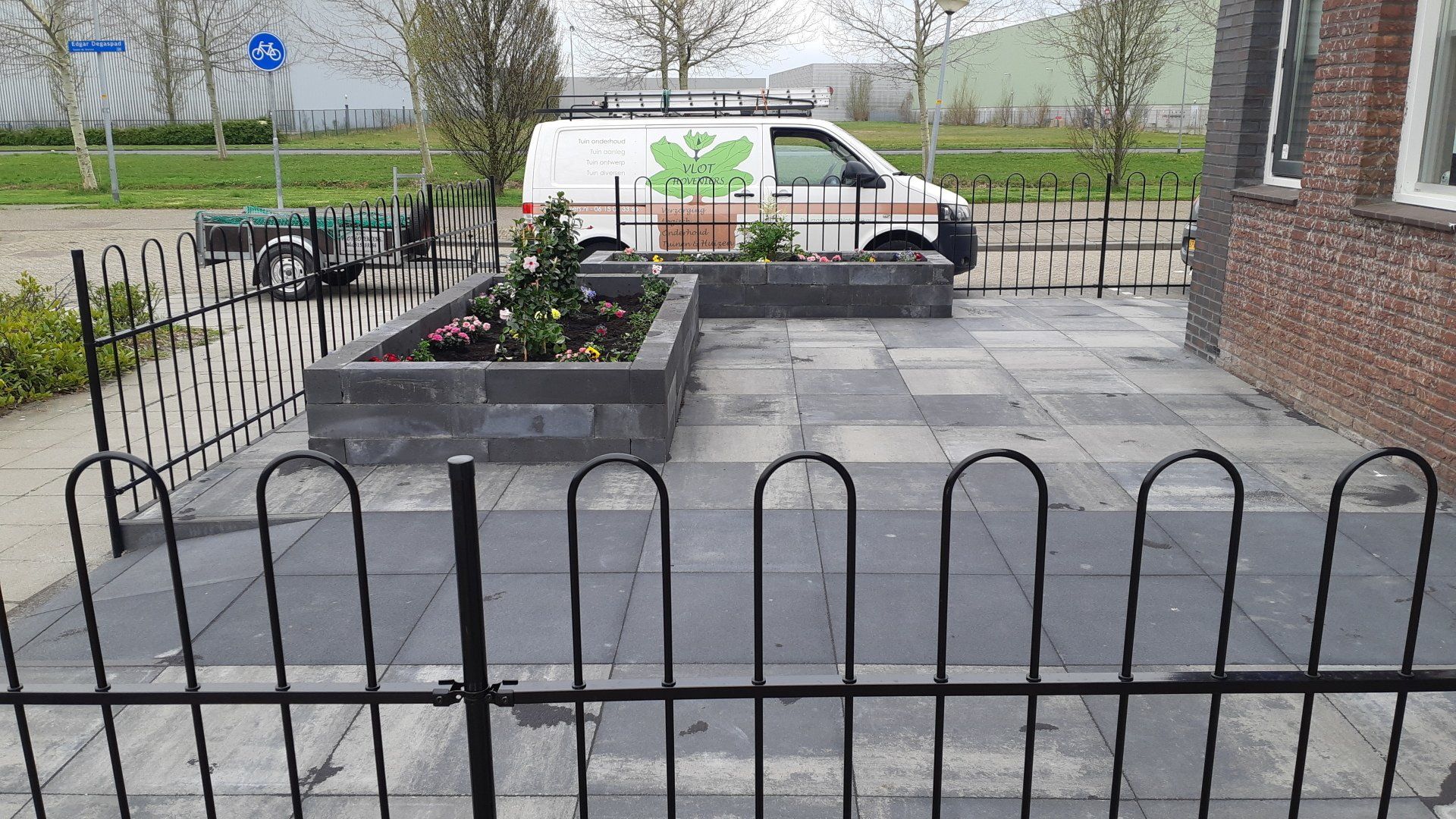 Kleine voortuin met verhoogde plantenbakken, een ijzeren hek en een geparkeerde witte bestelbus op een verharde oprit.