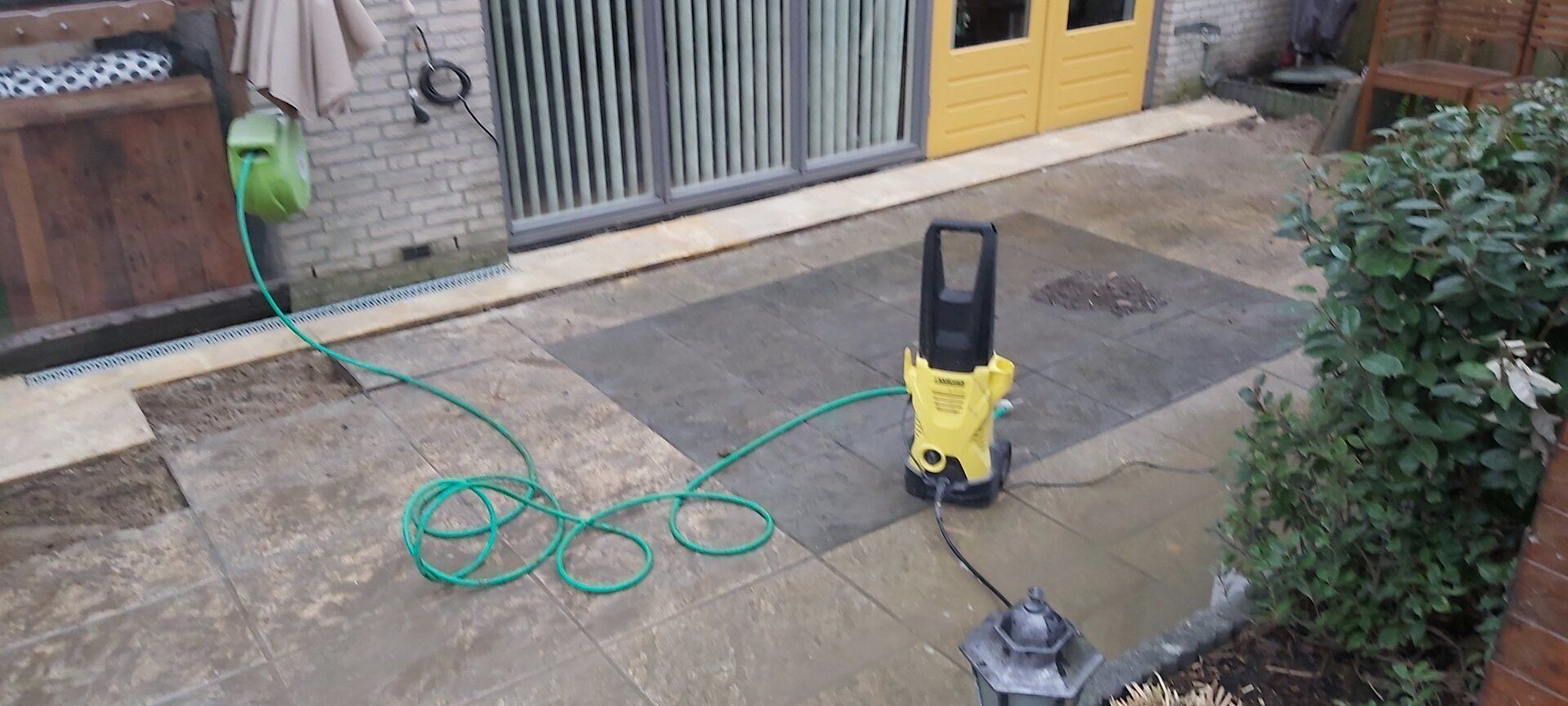 Gele hogedrukreiniger op een nat trottoir met een groene slang vlakbij een gebouwingang.