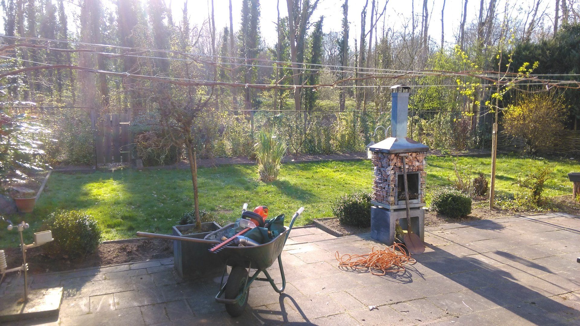 Terras in de achtertuin met kruiwagen, potplanten en een klein buitenfornuisje, te midden van bomen en zonlicht.