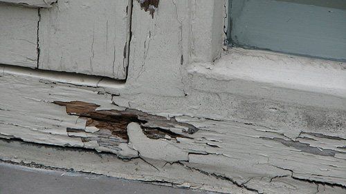 Afbladderende witte verf en rot hout op een buitenkozijn van een raam.