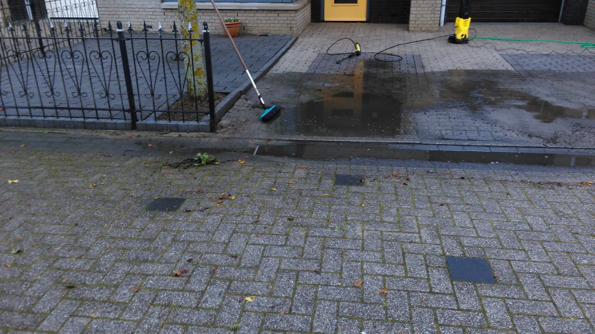 Een nat trottoir voor een gebouw, met een bezem die tegen een zwart hek leunt en plassen op de stoep.