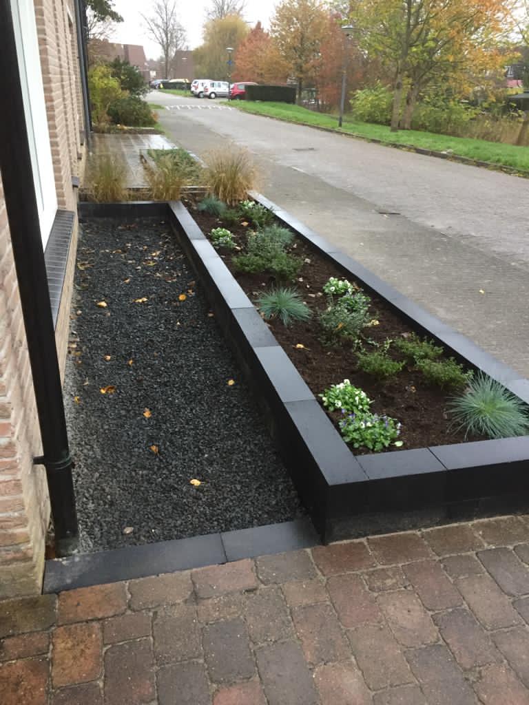 Voortuin met bakstenen pad en verhoogd, zwart omzoomd bloembed, grenzend aan een woonstraat.