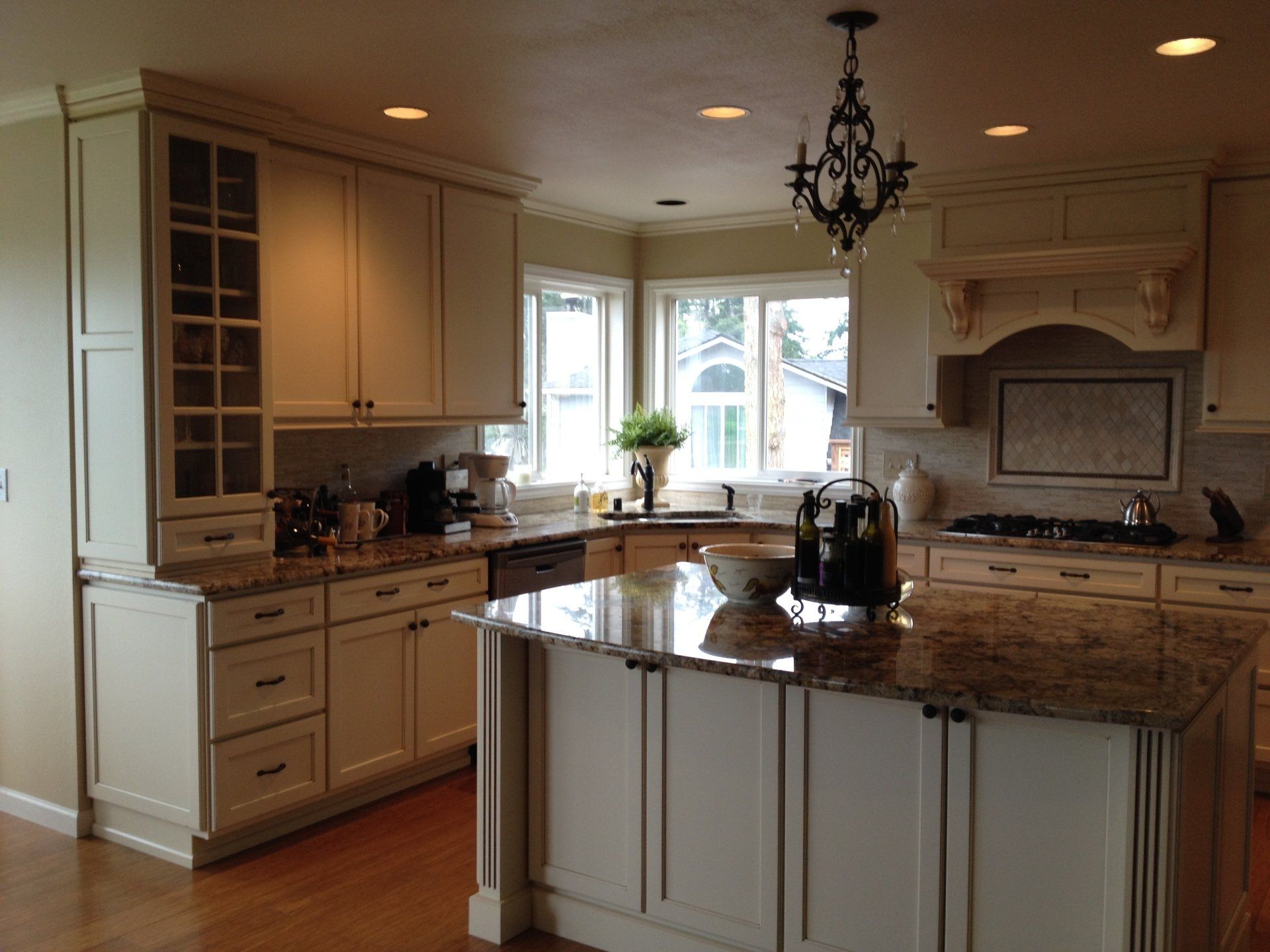 Bright kitchen with cream cabinets, granite island, chandelier, and sunlight through windows.