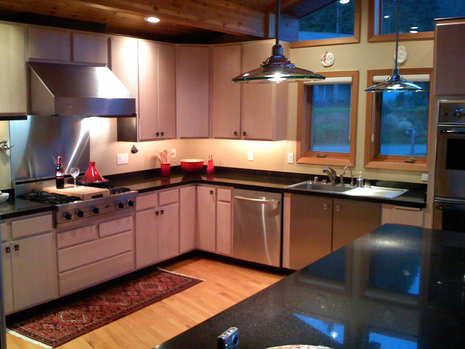 Modern kitchen with wood cabinets, stainless appliances, black countertops, and blue-lit windows.