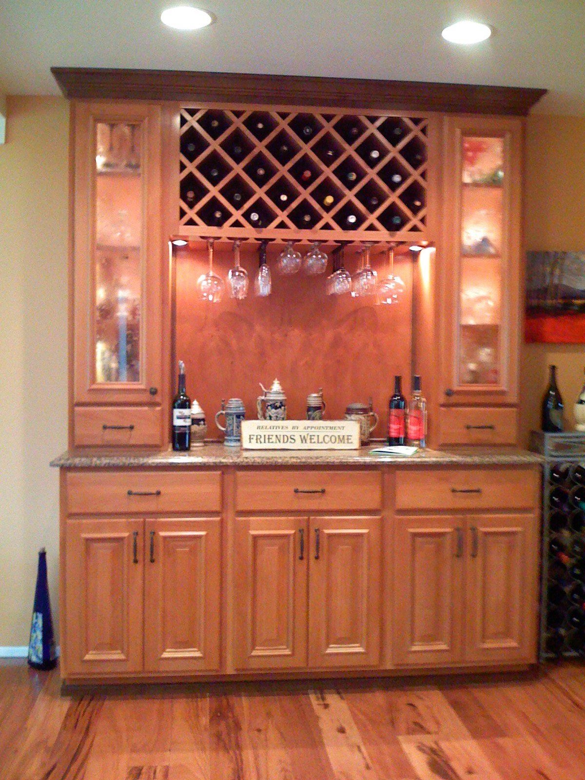 Wooden home bar cabinet with wine rack, glass shelves, and bottles against a wall, on hardwood floor.