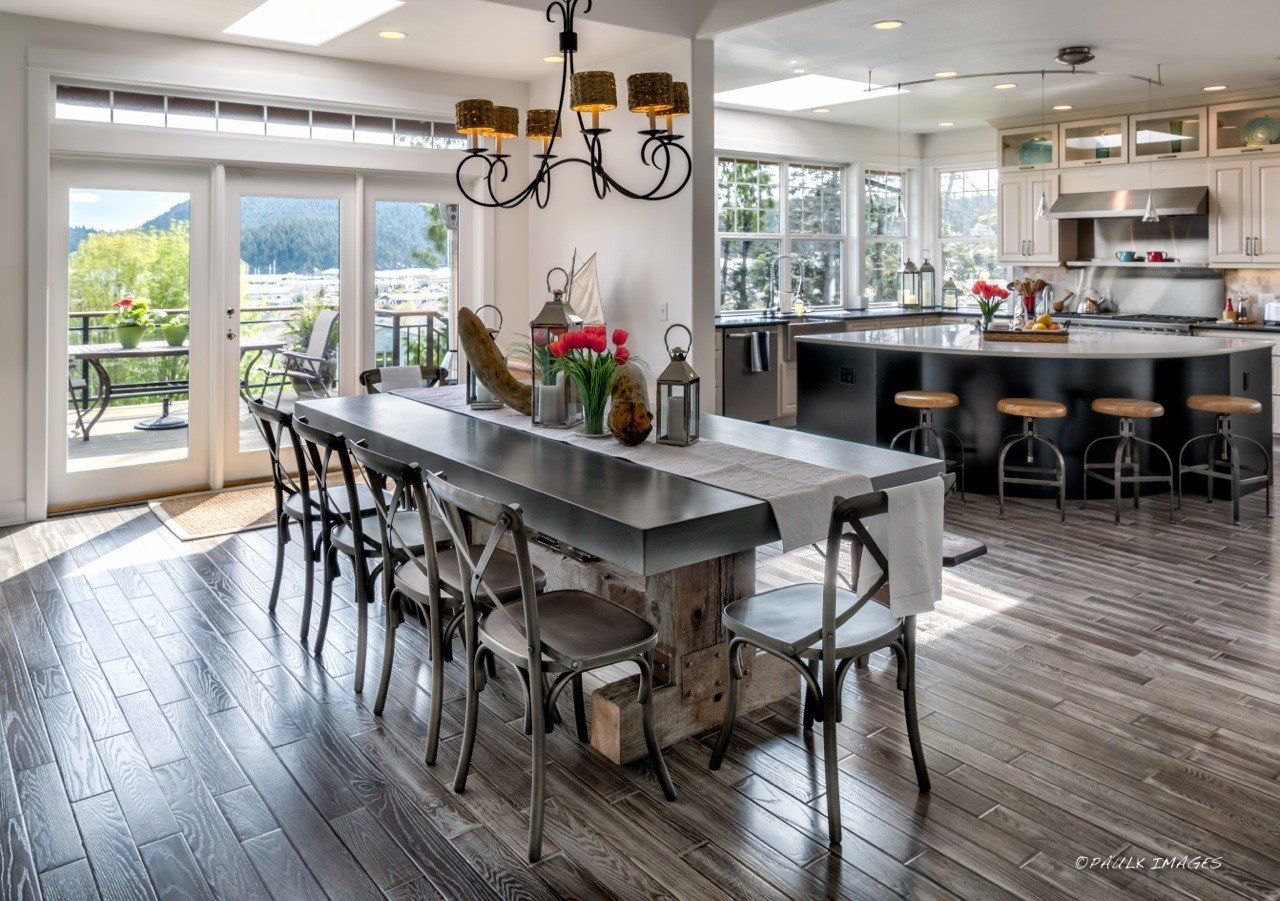 Bright open kitchen and dining area with long table, black stools, and large windows overlooking a patio.